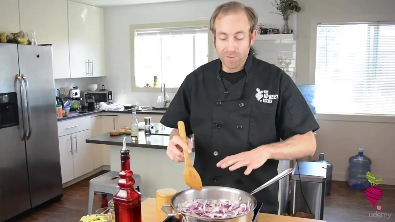 A chef in a black uniform stirs chopped red onions in a stainless steel pot using a wooden spoon in a bright kitchen.