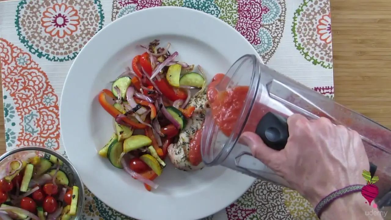 A hand pours red sauce from a blender onto a plate of cooked chicken and mixed vegetables on a patterned tablecloth.