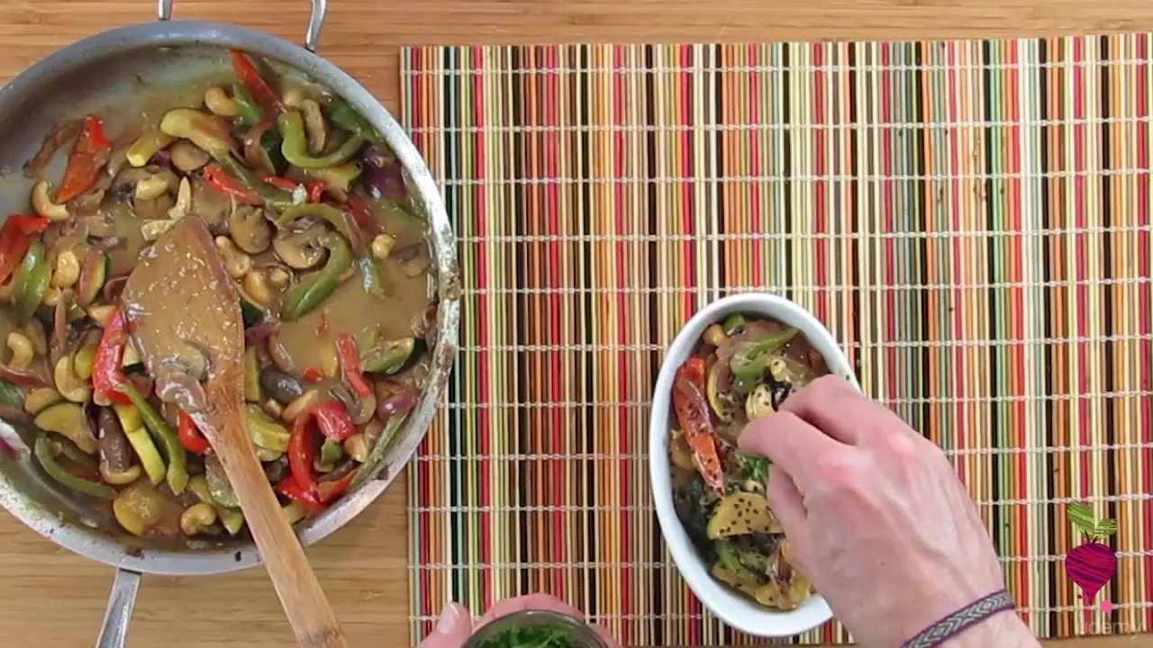 Overhead view of a pan with sautéed peppers, mushrooms, and cashews in brown sauce beside a white bowl where a hand adds ingredients.