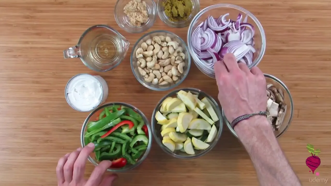 Overhead view of glass bowls holding chopped onions, cashews, peppers, lemon wedges, and mushrooms on a wooden surface with hands arranging them